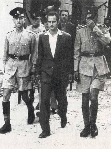 Ο Καραολής οδηγείται στο δικαστήριο. Θα καταδικασθεί σε θάνατο και θα εκτελεσθεί από τους Άγγλους δι’ απαγχονισμού στις 10 Μαΐου 1956. Δημοσιεύεται στην εφημερίδα Καθημερινή. Σάββατο 24-Κυριακή 25 Μαΐου 2014.